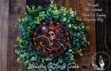 A wreath featuring a colorful design of a skeleton wearing a headdress, surrounded by green foliage and flowers. The wreath is displayed on a wooden surface.