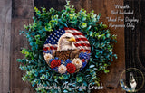 a wreath with an American flag design, surrounded by greenery and flowers. The wreath is displayed on a wooden surface, and there is a small dog in the bottom right corner of the image.