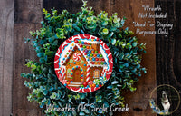 A wreath made of green foliage and featuring a gingerbread house design, with a caption indicating that the wreath is not included in a display and is used for purposes only.