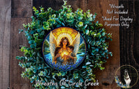 a wreath with a central image of an angel holding a child, surrounded by green foliage. The wreath is displayed on a wooden surface, and there is a small dog in the bottom right corner.