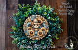 a wreath with a scarecrow design, surrounded by various green plants and flowers. The wreath is displayed on a wooden surface, and there is a small dog in the bottom right corner of the image.