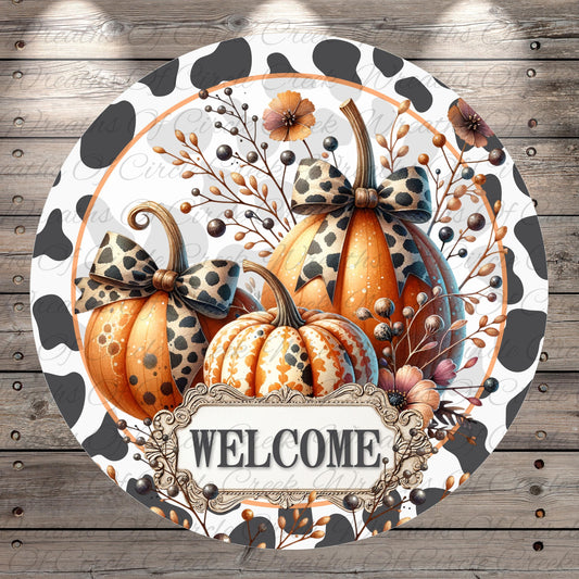 A circular welcome sign featuring three decorative pumpkins with bows, surrounded by fall foliage and flowers. The sign has a black and white spotted pattern and the word "WELCOME" is displayed in the center.
