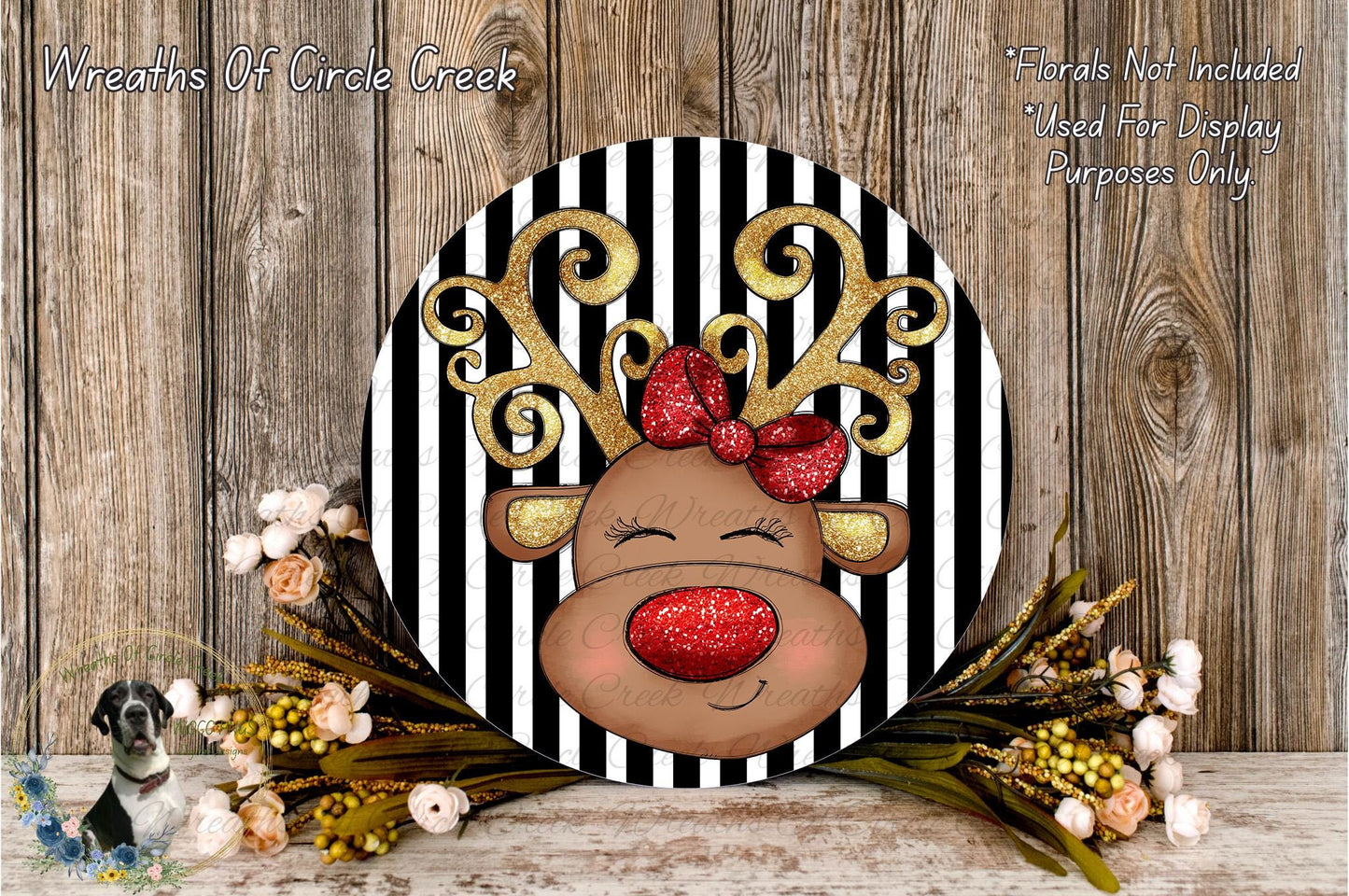 A decorative wreath featuring a red-nosed reindeer with a bow, surrounded by flowers and placed on a wooden surface.