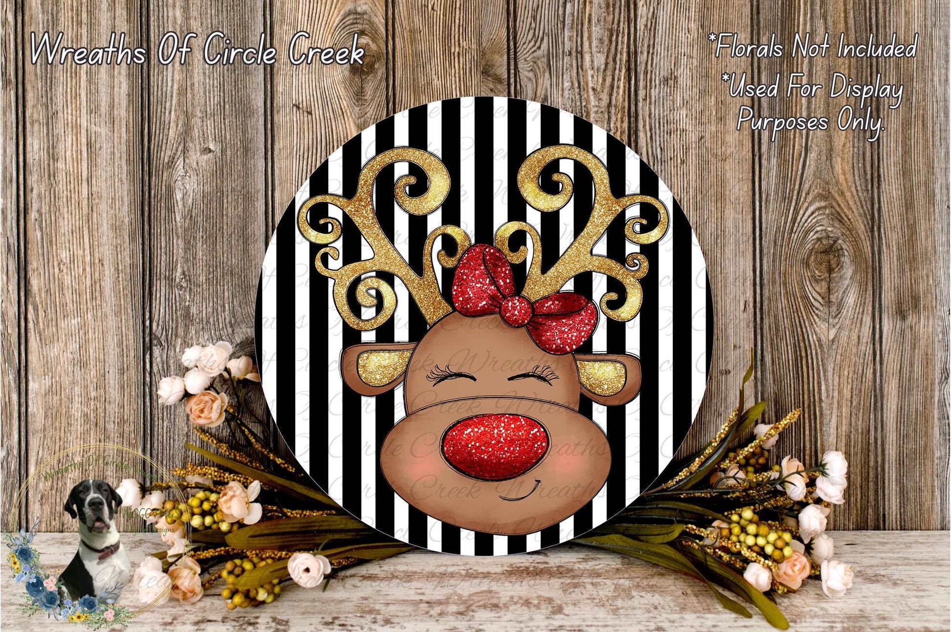 A decorative wreath featuring a red-nosed reindeer with a bow, surrounded by flowers and placed on a wooden surface.