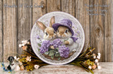 A decorative plate featuring two rabbits wearing purple hats, surrounded by purple flowers and greenery, placed on a wooden surface.