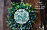 A wreath made of green leaves and a circular sign with the text "Merry Christmas" on it, placed on a wooden surface.