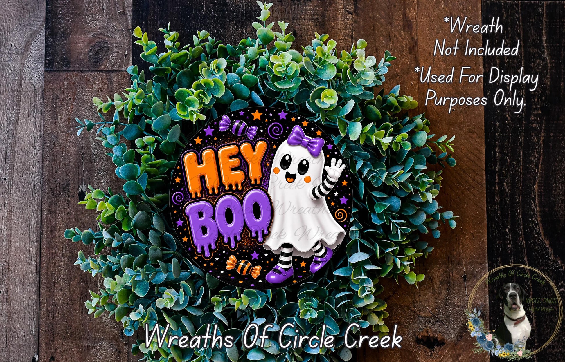 a Halloween-themed wreath made of green foliage and a purple ghost with the text "Hey Boo" on it. The wreath is displayed on a wooden surface, and there is a small dog in the bottom right corner of the image.