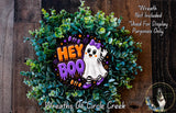 a Halloween-themed wreath made of green foliage and a purple ghost with the text "Hey Boo" on it. The wreath is displayed on a wooden surface, and there is a small dog in the bottom right corner of the image.