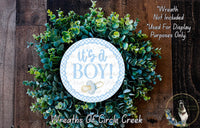 a wreath with the text "It's a BOY!" displayed on it, surrounded by green foliage. The wreath is placed on a wooden surface, and there is a small dog visible in the bottom right corner of the image.