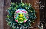 a wreath with a frog sitting on a pond surrounded by greenery. The wreath is placed on a wooden surface, and there is a text overlay that reads "Welcome Wreath Not Included * Used For Display Purposes Only" and "Wreaths of Circle Creek".