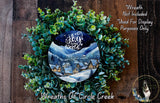 A wreath made of green leaves and a circular design featuring a winter village scene, with a blue sky and stars above.