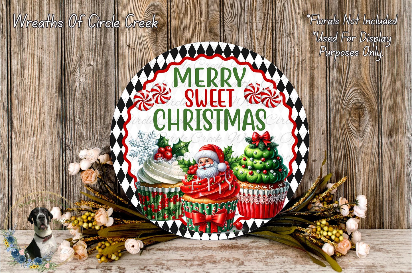 A festive Christmas-themed wreath with a Santa Claus figure, cupcakes, and other holiday decorations. The wreath is displayed on a wooden surface, surrounded by flowers.