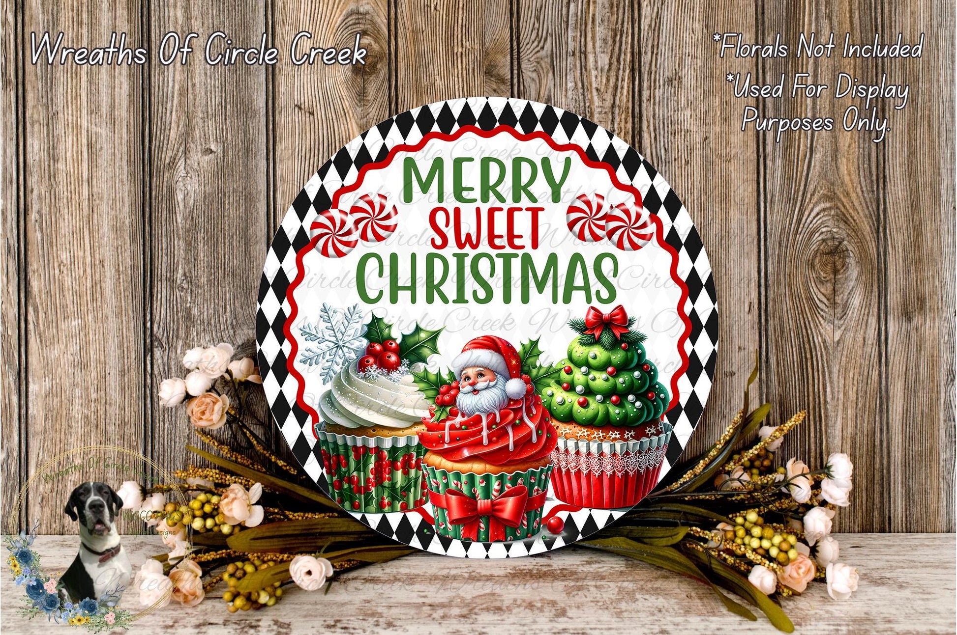 A festive Christmas-themed wreath with a Santa Claus figure, cupcakes, and other holiday decorations. The wreath is displayed on a wooden surface, surrounded by flowers.