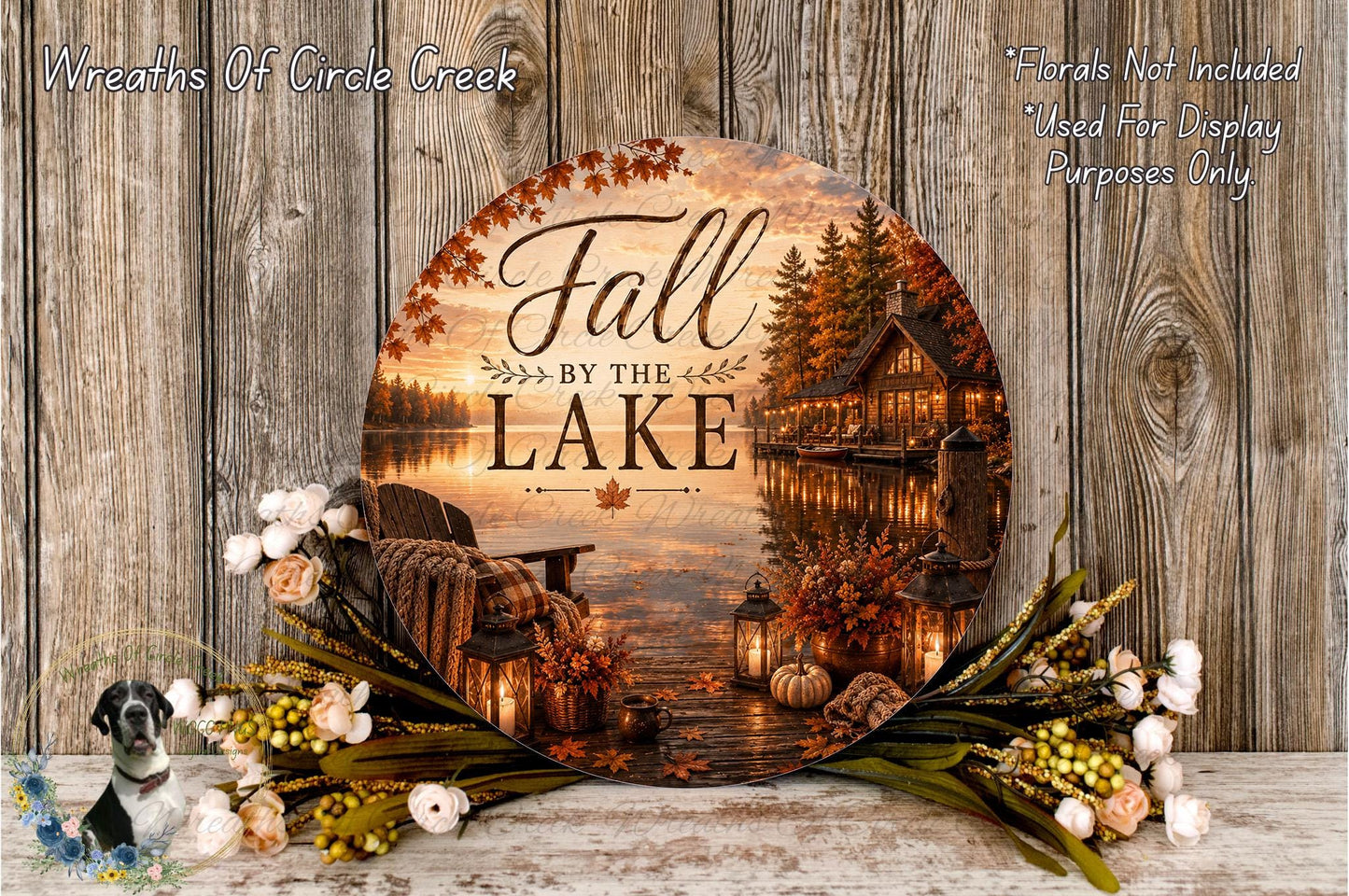 a round wooden sign with the text "Fall by the Lake" and a scenic image of a lake surrounded by autumn foliage, with a cabin and lanterns in the background. The sign is displayed on a wooden surface, and there are flowers and greenery surrounding it.