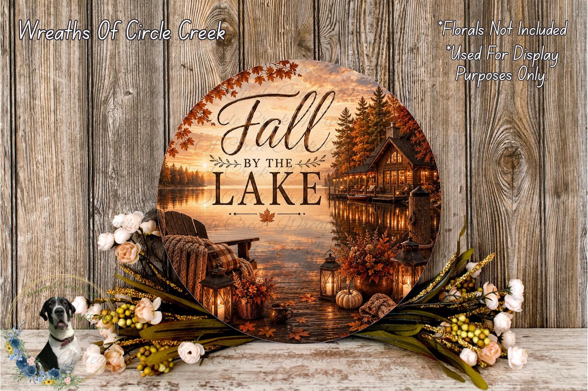 a round wooden sign with the text "Fall by the Lake" and a scenic image of a lake surrounded by autumn foliage, with a cabin and lanterns in the background. The sign is displayed on a wooden surface, and there are flowers and greenery surrounding it.