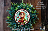 A wreath made of green leaves and featuring a cartoon elf wearing a red and white striped outfit.