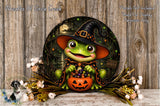 A whimsical Halloween-themed wreath featuring a green frog dressed as a witch, holding a carved pumpkin and surrounded by flowers and greenery.
