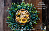 A wreath made of green foliage and flowers, with a bee and honeycomb design, and the text "Bee Happy" on it. The wreath is displayed on a wooden surface, and there is a small dog in the bottom right corner.