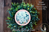a wreath made of green leaves and a circular sign with the text "Sea You at the Beach" on it, placed on a wooden surface.