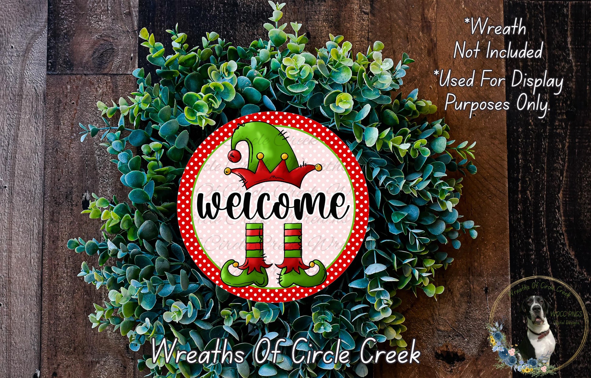 a festive wreath with a red and green elf sign, surrounded by greenery. The wreath is displayed on a wooden surface, and there is a small dog in the bottom right corner of the image.