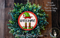 a festive wreath with a red and green elf sign, surrounded by greenery. The wreath is displayed on a wooden surface, and there is a small dog in the bottom right corner of the image.
