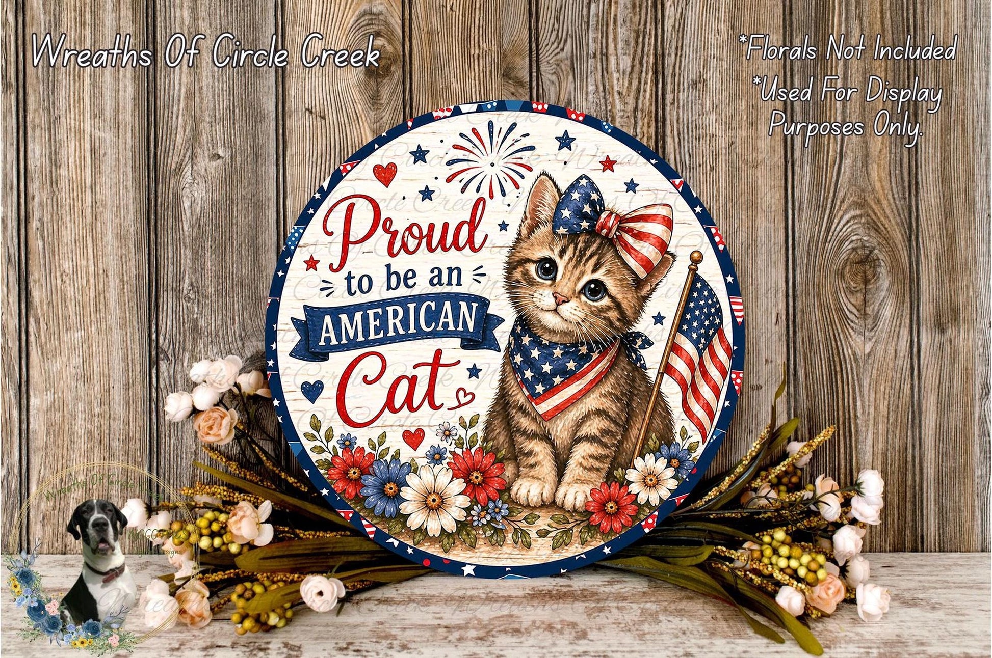 A patriotic-themed wreath with a cat wearing an American flag and ribbon, surrounded by flowers and a wooden background.
