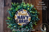 a wreath made of green leaves and a circular emblem with the text "Proud NAVY BRAT" on it. The wreath is displayed on a wooden surface, and there is a small dog in the bottom right corner of the image.