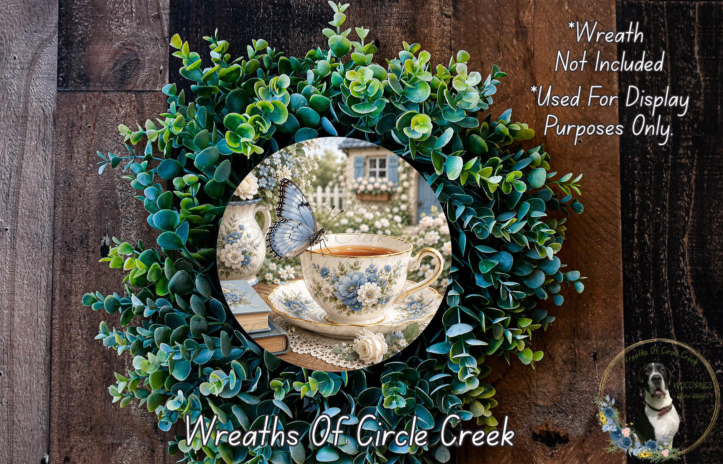 A wreath made of green leaves and flowers, with a teacup and a butterfly resting on it. The wreath is displayed on a wooden surface, and there is a text overlay that reads "Wreath Not Included * Used For Display Purposes Only".