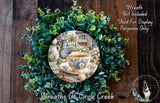 A wreath made of green leaves and flowers, with a teacup and a butterfly resting on it. The wreath is displayed on a wooden surface, and there is a text overlay that reads "Wreath Not Included * Used For Display Purposes Only".