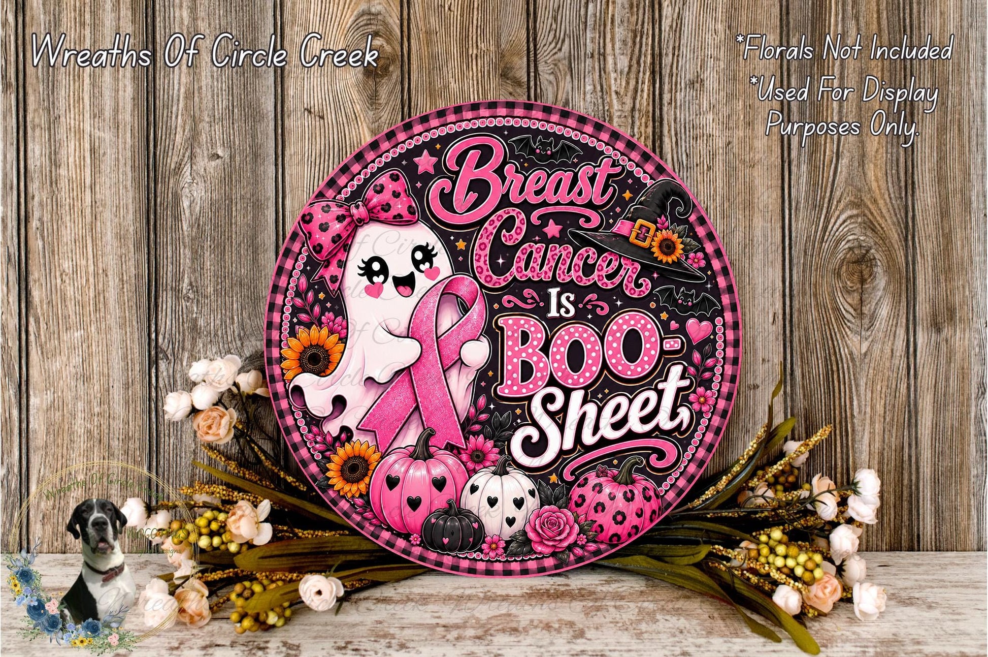 A round sign with a pink and black design, featuring a ghost holding a pink ribbon and the text "Breast Cancer is Boo- Sheet". The sign is surrounded by flowers and placed on a wooden surface.
