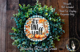 a wreath made of various green and orange leaves, with a central circular design that says "It's Fall Yall". The wreath is displayed on a wooden surface, and there is a small dog in the bottom right corner of the image.