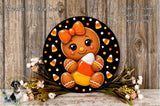 A gingerbread girl with a bow and candy cane is displayed on a black circular plaque, surrounded by a floral arrangement and candy canes.