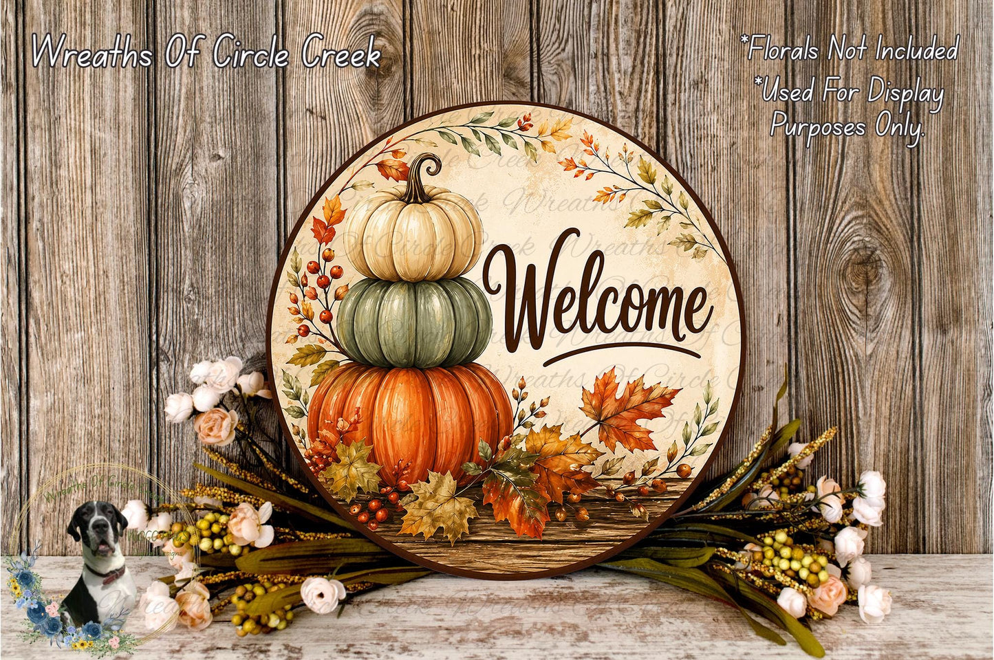 A decorative welcome sign with a stack of three pumpkins and autumn leaves, surrounded by flowers and placed on a wooden surface.
