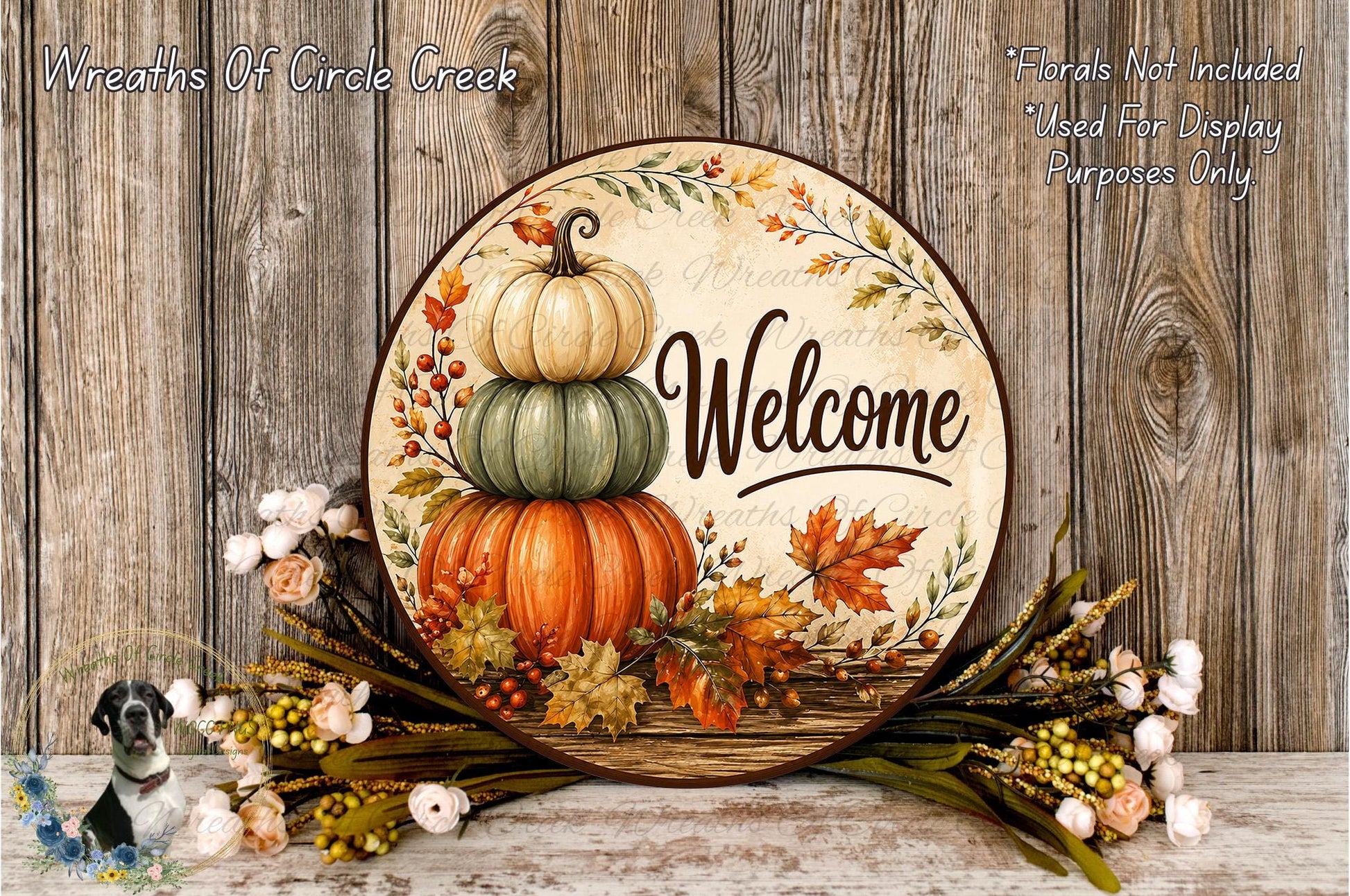 A decorative welcome sign with a stack of three pumpkins and autumn leaves, surrounded by flowers and placed on a wooden surface.
