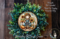 a wreath with a scarecrow and sunflowers, surrounded by greenery. The wreath is displayed on a wooden surface, and there is a small dog in the bottom right corner.