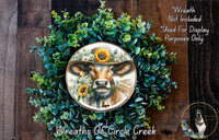 a wreath with a cow wearing a hat and surrounded by greenery, including sunflowers. The wreath is displayed on a wooden surface, and there is a small dog in the bottom right corner.