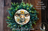 a wreath with a cow wearing a hat and surrounded by greenery, including sunflowers. The wreath is displayed on a wooden surface, and there is a small dog in the bottom right corner.