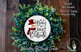 A wreath made of green plants and a snowman decoration, with the text "Baby is cold outside" displayed on it.