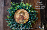 a wreath with a scarecrow design, surrounded by a lush green plant. The wreath is placed on a wooden surface, and there is a small dog visible in the bottom right corner of the image.