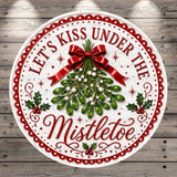 A decorative circular sign with the text "Let's Kiss Under The Mistletoe" and a decorative ribbon, surrounded by a festive design with holly leaves, berries, and pine cones.
