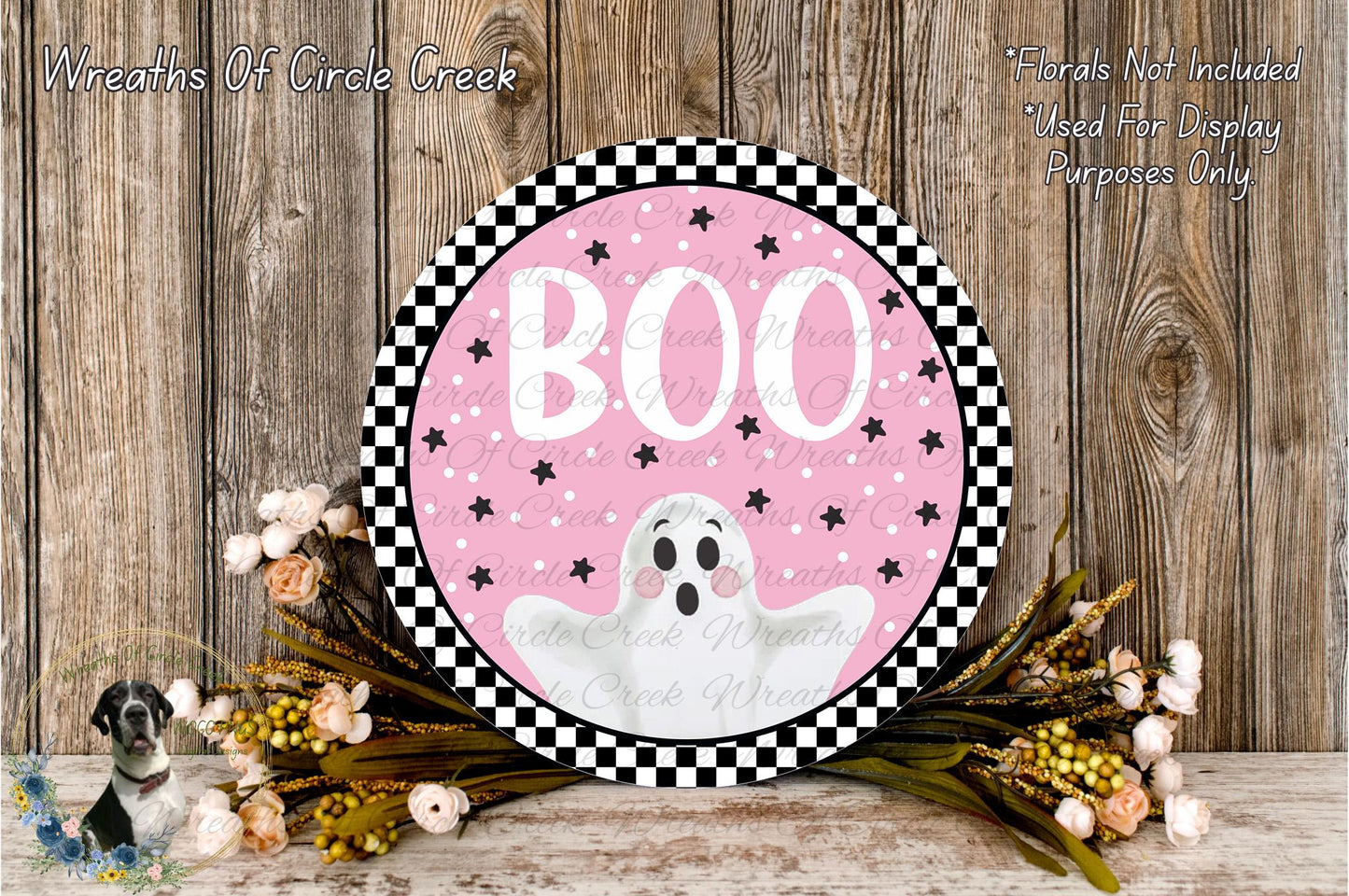 a Halloween-themed decoration with a pink background, black and white checkered border, and a ghost illustration. The decoration is surrounded by a floral arrangement and placed on a wooden surface.