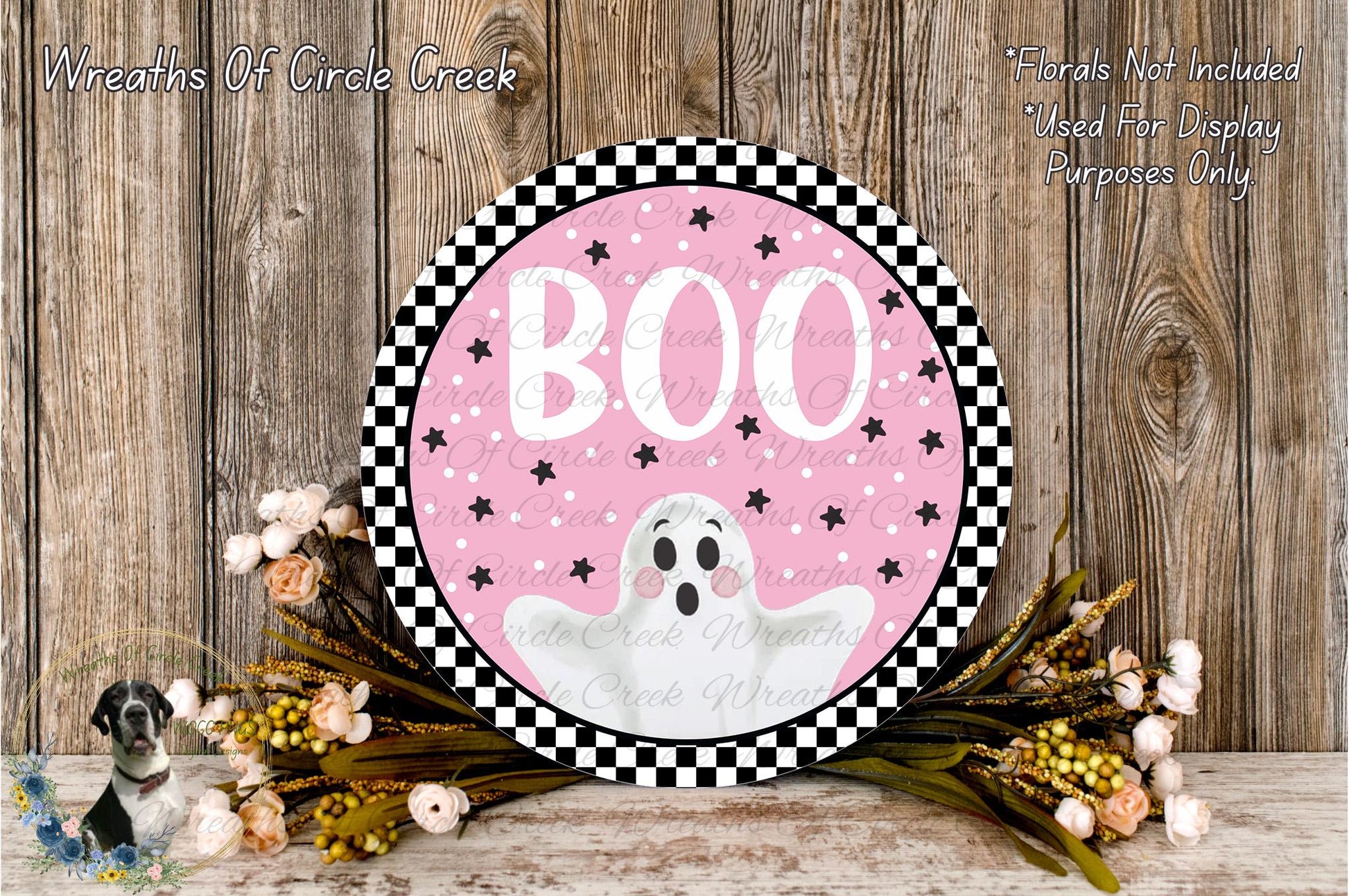 a Halloween-themed decoration with a pink background, black and white checkered border, and a ghost illustration. The decoration is surrounded by a floral arrangement and placed on a wooden surface.