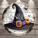 A witch's hat with the text "The Witch is in" and a pumpkin, along with a spider web and bats, is displayed on a wooden surface.