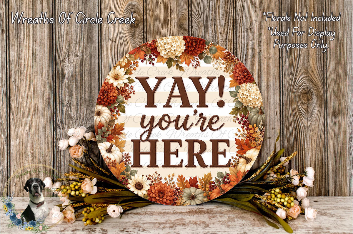 a decorative sign with the text "YAY! you're here" surrounded by a floral arrangement on a wooden surface.