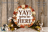 a decorative sign with the text "YAY! you're here" surrounded by a floral arrangement on a wooden surface.