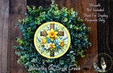 a wreath made of various green plants and flowers, including a cross at the center. The wreath is displayed on a wooden surface, and there is a small dog in the bottom right corner of the image.