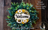 a wreath with a welcoming sign, surrounded by a lush green wreath with white and yellow flowers, and a dog in the background.