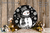 A snowman wearing a scarf and hat, surrounded by flowers and placed on a wooden surface.