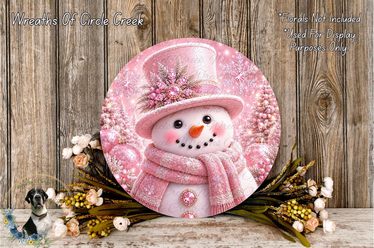 A round, decorative snowman with a pink top hat and scarf, surrounded by pink flowers and greenery, set against a wooden background.