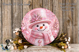 A round, decorative snowman with a pink top hat and scarf, surrounded by pink flowers and greenery, set against a wooden background.